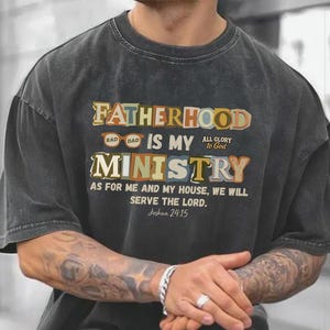 May include: A dark gray t-shirt with the text "FATHERHOOD IS MY MINISTRY" in a colorful, vintage-style design. The shirt also features the Bible verse Joshua 24:15. The shirt is a relaxed fit.