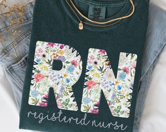 May include: A dark teal t-shirt with the letters "RN" in a floral pattern, with the words "registered nurse" below. The shirt is folded on top of a pair of blue jeans, with a gold necklace and dried flowers. The shirt is labeled "Blue Spruce".