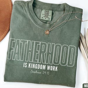 May include: A moss green t-shirt with the words "FATHERHOOD IS KINGDOM WORK" in white, with "Joshua 24:15" below. A gold necklace and a brown hat are also visible. The shirt is a Comfort Colors brand.
