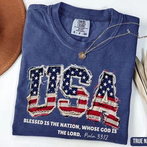 May include: True navy blue t-shirt with the word "USA" in a patriotic design featuring the American flag. The shirt includes the text "Blessed is the nation, whose God is the Lord. Psalm 33:12". A gold necklace rests on the shirt.