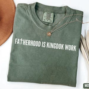 May include: A moss green t-shirt with the text "FATHERHOOD IS KINGDOM WORK" in white. A gold necklace with a pendant is draped on the shirt. A brown hat and dried leaves are in the background.
