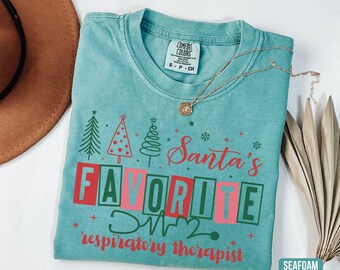 May include: Seafoam green t-shirt with the text "Santa's FAVORITE respiratory therapist" in red and green. The design includes Christmas trees, snowflakes, and a stethoscope. A gold necklace is draped across the shirt.