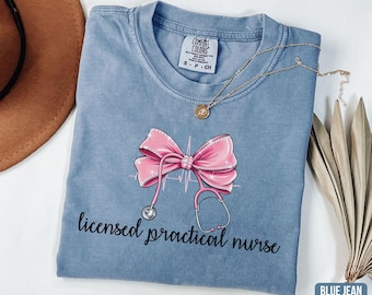 May include: Blue jean colored t-shirt with a pink bow and stethoscope graphic. The text "licensed practical nurse" is printed below the graphic. A gold necklace is draped across the shirt.