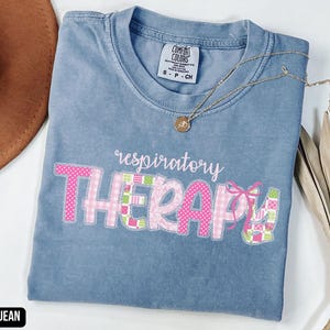 May include: Blue jean colored t-shirt with the words "respiratory THERAPY" in pink and green patchwork letters. A gold necklace with a pendant is draped across the shirt. The shirt is folded on a white surface.