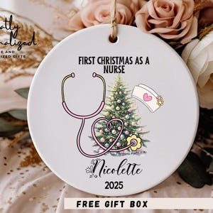 May include: A round white ceramic ornament featuring a graphic of a Christmas tree, a pink stethoscope shaped into a heart, and a nurse cap. The text reads FIRST CHRISTMAS AS A NURSE, the name Nicolette, and the year 2025 in black lettering.