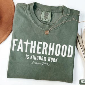 May include: A moss green t-shirt with the words "FATHERHOOD IS KINGDOM WORK" printed in white. The text "Joshua 24:15" is below the main text. A gold necklace with a pendant rests on the shirt.