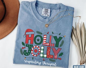 May include: Blue jean Comfort Colors t-shirt with a festive "HOLLY JOLLY" design in red, green, and white, accented with a Santa hat and Christmas lights. The text "respiratory therapist" is below the design. A gold necklace rests on the shirt.