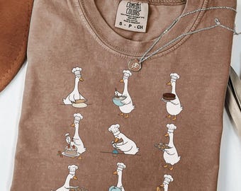 May include: A brown t-shirt featuring a graphic print of nine white geese wearing chef hats and performing various baking tasks. The shirt has a Comfort Colors brand label at the inner neckline and a silver-toned necklace resting on the fabric.