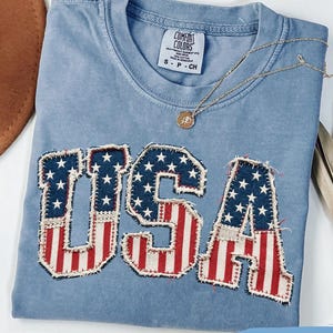 Puede incluir: Una camiseta azul vaquera con la palabra "USA" aplicada en la parte delantera con un diseño patriótico. Las letras presentan la bandera estadounidense con rayas rojas y blancas y estrellas azules. Un collar de oro descansa sobre la camisa.
