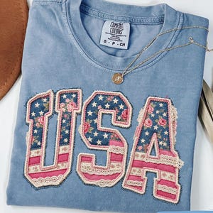 Puede incluir: Una camiseta Comfort Colors azul vaquero con la palabra "USA" aplicada en la parte delantera. Las letras presentan un diseño floral y estrellado con detalles en rosa y encaje. Un collar de oro con un colgante descansa sobre la camiseta.