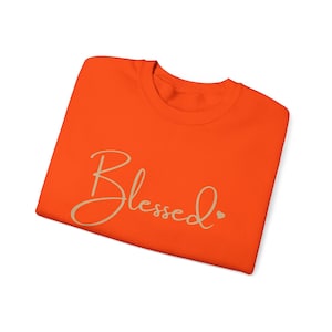 May include: Orange t-shirt with the word "Blessed" written in a cursive font in beige with a small heart.