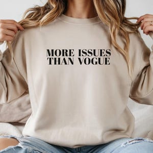 May include: Beige sweatshirt with the text "MORE ISSUES THAN VOGUE" in black, bold font. The sweatshirt is a neutral color and has a crew neck. The image shows the sweatshirt on a person.