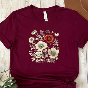 May include: Burgundy t-shirt featuring a floral design with white, red, and green flowers. The graphic is centered on the front of the shirt. The shirt has rolled-up sleeves and a crew neck.