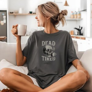 May include: Dark gray t-shirt with the words "DEAD TIRED" above a skull graphic and a coffee cup. The shirt is a relaxed fit and appears to be made of a soft material. The design is in a distressed, vintage style.