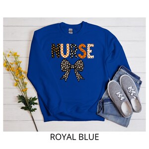 May include: Royal blue sweatshirt with the word "NURSE" in orange and black letters. A black and white bow is below the word. The sweatshirt is paired with blue jeans and gray sneakers. The text "ROYAL BLUE" is at the bottom.