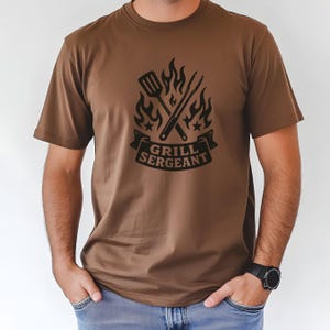 May include: Brown t-shirt with a black graphic featuring a spatula and tongs crossed over flames, with the text "GRILL SERGEANT" on a banner. The shirt is short-sleeved and has a crew neck.