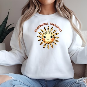 May include: White sweatshirt featuring a smiling sun graphic with black spots and the text "BEHAVIORAL THERAPIST" in brown. The sun has a yellow and orange color scheme. The sweatshirt is a casual style.