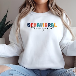 Könnte beinhalten: Weißes Sweatshirt mit dem Aufdruck "BEHAVIORAL therapist". Das Wort "BEHAVIORAL" ist in Regenbogenfarben gehalten, und "therapist" ist in schwarzer Schrift geschrieben. Das Sweatshirt hat einen lässigen Stil.