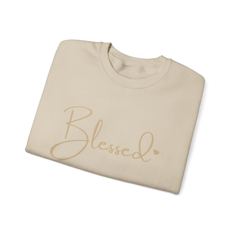 May include: A beige sweatshirt with the word "Blessed" written in a cursive font in gold. A small heart symbol is next to the word.
