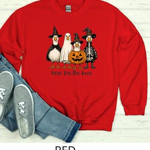 May include: A red sweatshirt featuring a Halloween-themed graphic. The graphic includes four cartoon ducks dressed in Halloween costumes: a witch, a ghost, a jack-o'-lantern, and a skeleton. The text "here for the boos" is printed below the ducks.