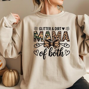 May include: Beige sweatshirt with the text "Glitter & Dirt Mama of both" in black, camouflage, and leopard print. A bow graphic is centered below the word "Mama". The sweatshirt has long sleeves.