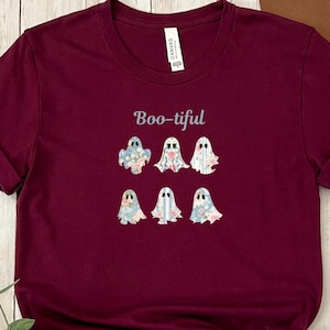 May include: A maroon t-shirt featuring the word "Boo-tiful" above six ghost illustrations. Each ghost has a floral pattern in shades of blue, pink, and white. The shirt is made of a soft material.