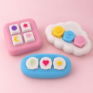 Op de afbeelding: Drie fidget-speeltjes met tactiele knoppen. Een roze vierkante basis met vier witte vierkante toetsen met een paarse bloem, rode ster, gele maan en roze hart. Een witte wolk-vormige basis met drie ronde toetsen: gele ster, blauwe maan, roze hart.