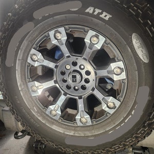 May include: Close-up of a black tire with a silver rim and the text "AT II" and "ALL-TERRAIN COMPOUND" on the side. The tire is on a jack and has a gray painted line around the sidewall.