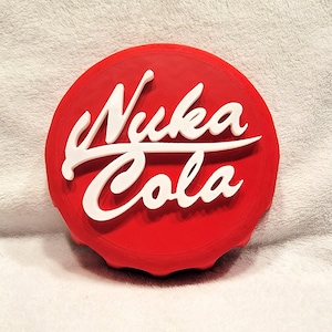 May include: A red Nuka Cola bottle cap with white raised lettering. The text "Nuka Cola" is prominently displayed across the center of the cap. The cap has a textured edge and sits on a white surface.