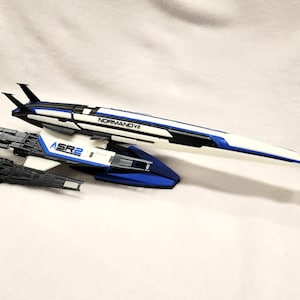 May include: A model of a spaceship, featuring a sleek design with white, blue, and black accents. The model has the text "NORMANDYA" and "ASR2" printed on it. The spaceship is set against a white background.