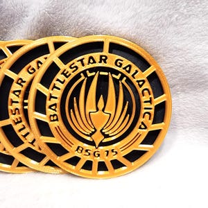 Puede incluir: Primer plano de varias fichas circulares doradas y negras. Las fichas presentan el texto "Battlestar Galactica" en un patrón circular alrededor de un emblema central. La parte inferior de la ficha dice "BSG 75". Las fichas están impresas en 3D.