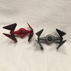 May include: Two TIE fighter starship models, one in a vibrant red finish and the other in a neutral gray finish. Both spacecraft feature a central spherical cockpit with a black circular window and two angular, black-trimmed solar array wings.