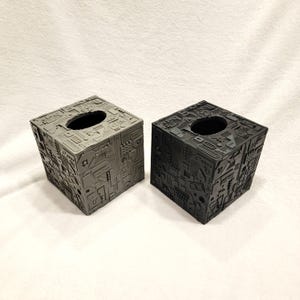 May include: Two square tissue box covers featuring a complex, mechanical, and geometric surface texture. One box is finished in a light gray, while the other is a matte black. Both feature a circular opening on the top for dispensing tissues.
