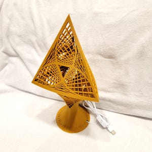 May include: A geometric, mustard-colored lamp with a pyramid-shaped shade. The shade features a lattice design, and the lamp has a round base and a USB power cord with an on/off switch. The lamp is approximately 10 inches tall.