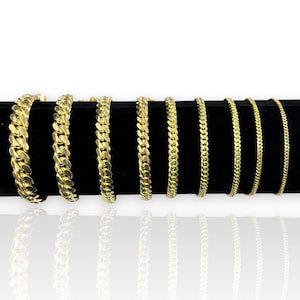 May include: A collection of gold-colored chain bracelets displayed on a black surface. The bracelets vary in thickness and style, showcasing a range of designs from thick curb chains to delicate link chains. The chains are reflective.
