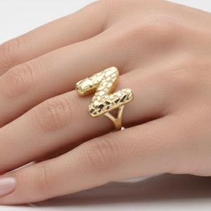 Puede incluir: Un anillo dorado con forma de la letra "Z" se muestra en una mano. El anillo tiene una superficie texturizada y está sobre un fondo blanco. El anillo es una joya.