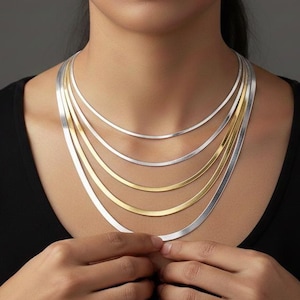 Puede incluir: Collar de varias hebras con cadenas planas en tonos plateados y dorados. El collar lo lleva una persona con un top negro. Cada cadena tiene una longitud diferente.