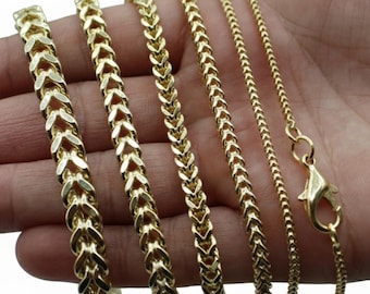 May include: Several gold-colored chain necklaces of varying styles are displayed. The chains include different link designs, such as a herringbone pattern and a thinner, more delicate chain. A clasp is visible on the rightmost chain.