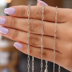 May include: Three delicate silver chain necklaces with a paperclip link design are displayed. The necklaces are held against a hand with light purple nail polish. The background is blurred, with soft, warm lighting.