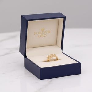 Puede incluir: Un anillo de oro con un dise&ntilde;o intrincado descansa dentro de una caja de joyer&iacute;a azul marino abierta. El interior crema lleva la inscripci&oacute;n "FOREVER ORO". El anillo, en el centro, destaca su detallado trabajo de joyer&iacute;a.