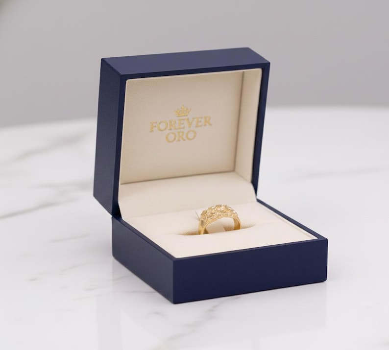 Puede incluir: Un anillo de oro con un dise&ntilde;o intrincado descansa dentro de una caja de joyer&iacute;a azul marino abierta. El interior crema lleva la inscripci&oacute;n "FOREVER ORO". El anillo, en el centro, destaca su detallado trabajo de joyer&iacute;a.
