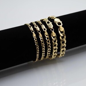 May include: A collection of gold-colored chain bracelets of varying thicknesses. The bracelets are displayed on a black cylindrical surface against a white background. Each bracelet features a secure clasp.