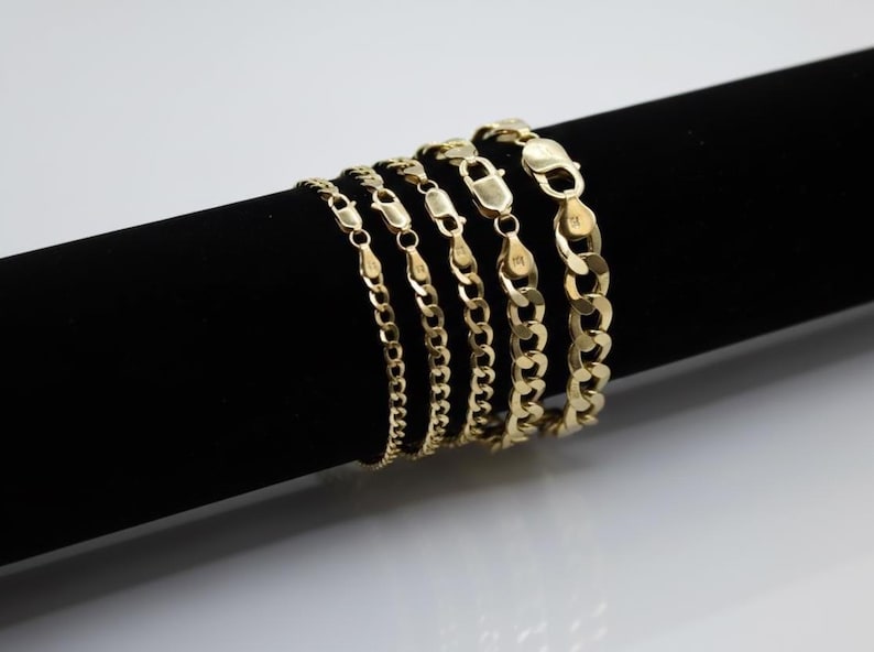 May include: A collection of gold-colored chain bracelets of varying thicknesses. The bracelets are displayed on a black cylindrical surface against a white background. Each bracelet features a secure clasp.