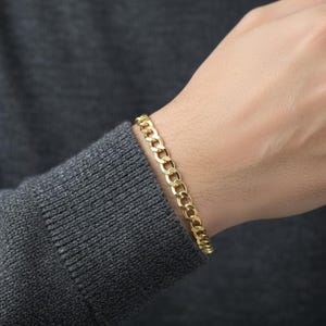 May include: A gold-colored curb chain bracelet is worn on a person's wrist. The bracelet is made of interlocking oval links. The person is wearing a gray sweater.
