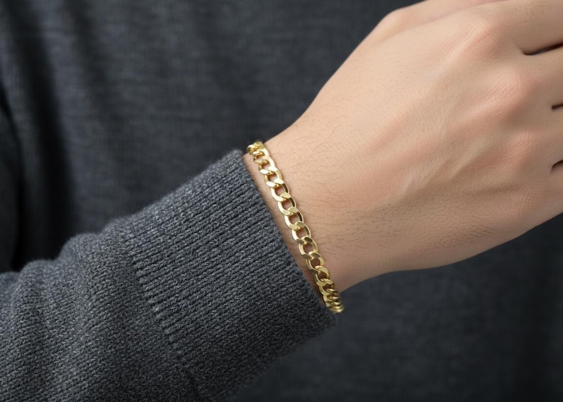 May include: A gold-colored curb chain bracelet is worn on a person's wrist. The bracelet is made of interlocking oval links. The person is wearing a gray sweater.