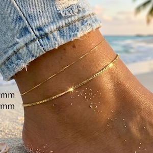 May include: Close-up of a foot wearing two delicate gold anklets, 0.8mm and 1.5mm thick, on a sandy beach. The anklets are thin and shiny, reflecting the sunlight. The person is wearing blue denim jeans.