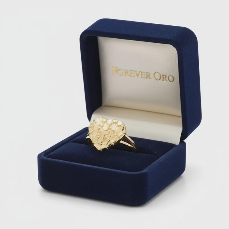 Puede incluir: Un anillo de oro con un dise&ntilde;o texturizado en forma de coraz&oacute;n, exhibido en una caja de anillo de terciopelo azul marino. El interior de la caja es blanco con el texto "FOREVER ORO" en letras doradas. El anillo tambi&eacute;n es de oro.