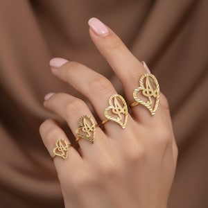 Puede incluir: Una mano luce cuatro anillos dorados en forma de corazón. Cada anillo presenta un delicado diseño calado con un motivo central. Los anillos varían ligeramente de tamaño, creando un efecto de capas. El fondo es marrón suave y borroso.