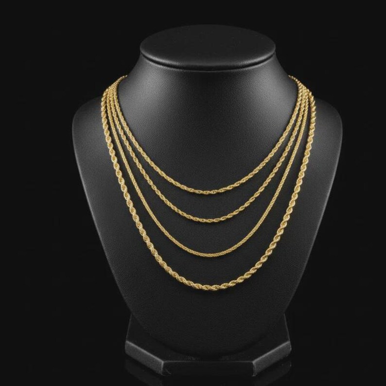 May include: Four gold-colored rope chain necklaces of varying thicknesses are displayed on a black jewelry bust. The necklaces are arranged in a layered style, showcasing different textures and designs. The background is a solid black, highlighting the jewelry.