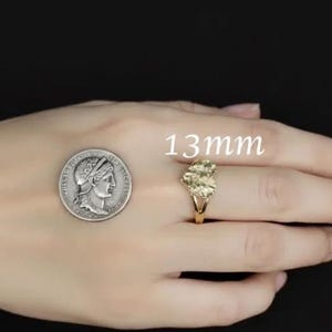 Puede incluir: Un anillo dorado con un grupo de piedras se muestra junto a una moneda. El anillo mide aproximadamente 1,3 cm. El anillo est&aacute; en un dedo. El fondo es negro.
